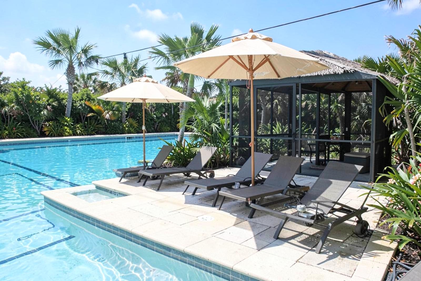 Pool deck with lounge chairs, umbrellas, a screened shade structure and palms. outdoor pool community amenity