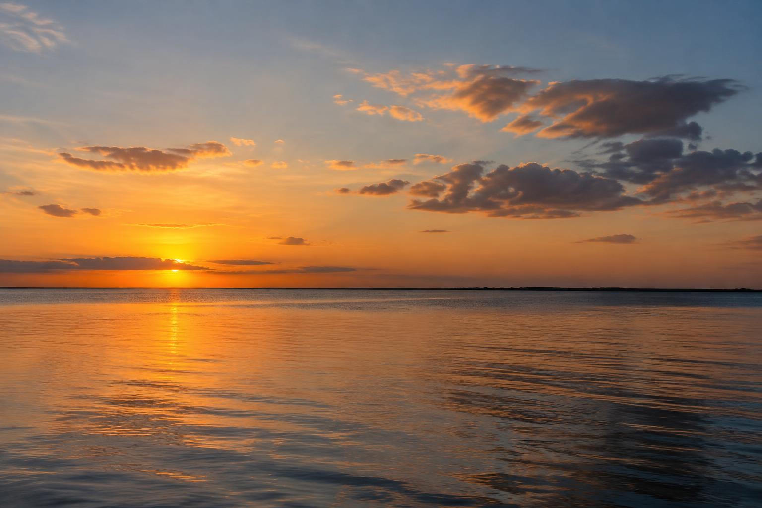 Calm coastal water beneath a warm sunset sky with gentle ripples and a low, unbroken horizon.