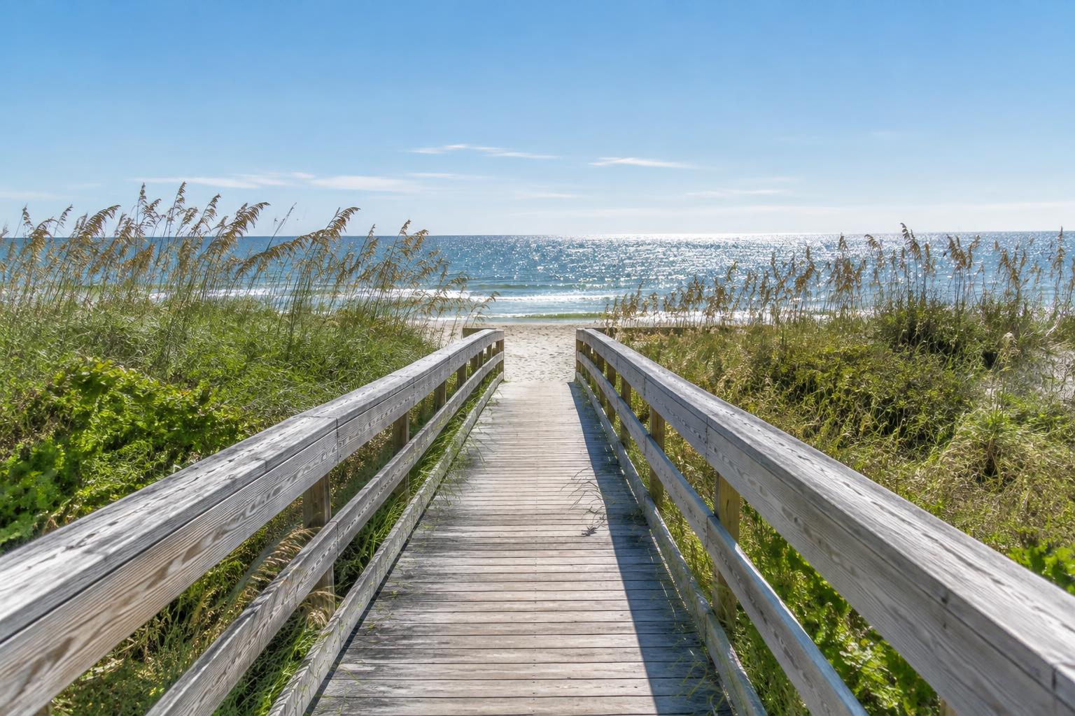 Wooden coastal boardwalk crossing sea oats and dune vegetation toward bright blue water.